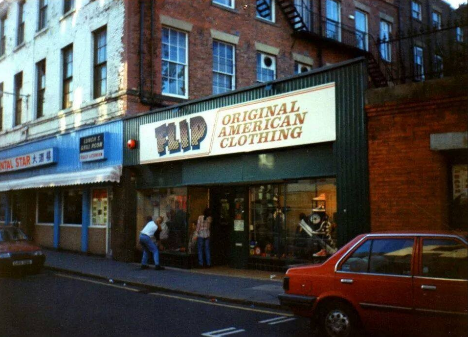 Storefront with a sign advertising 'Flip Original American Clothing' on a city street with a few people in front and cars parked along the curb.