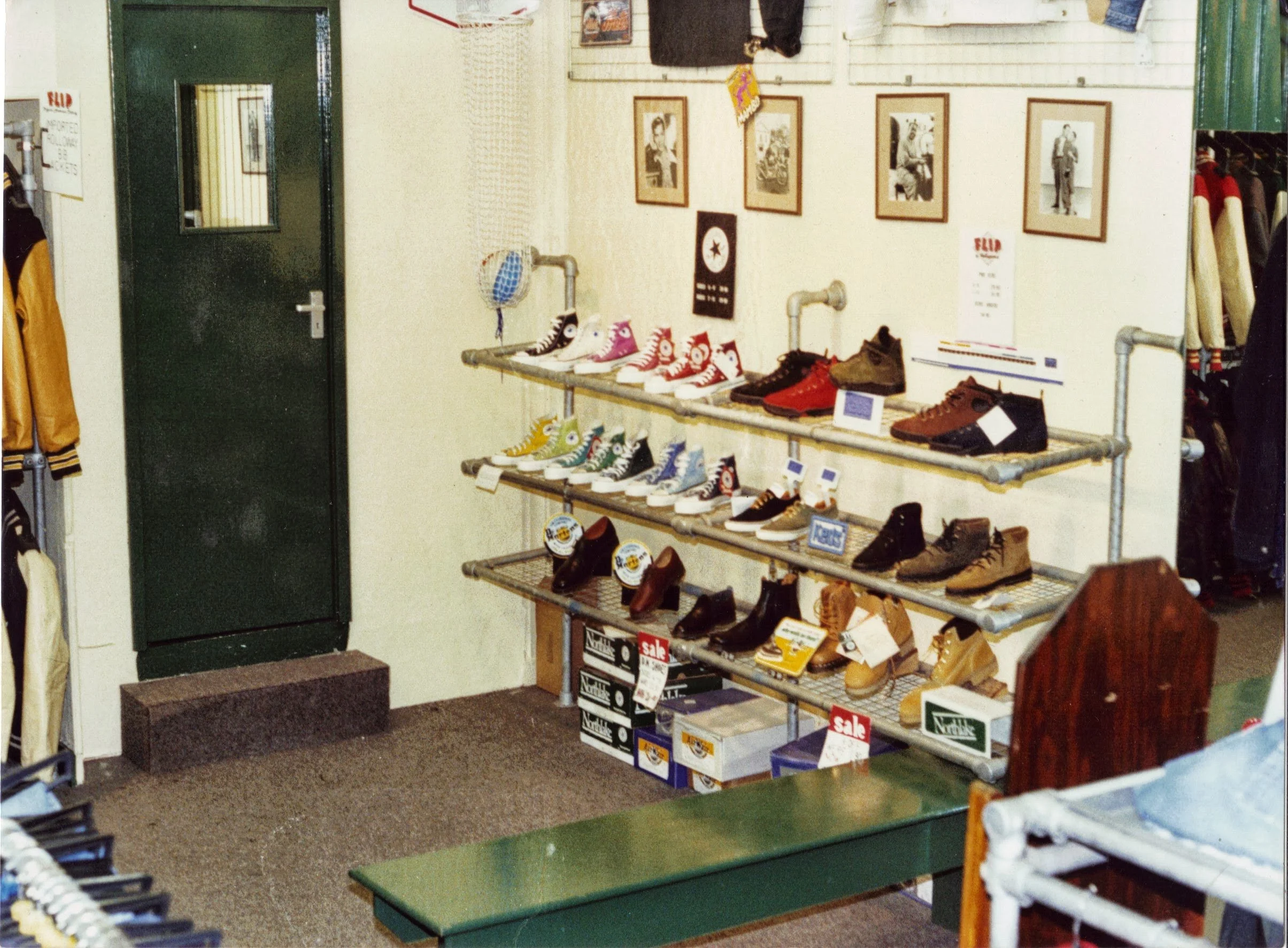 Inside a shoe store with a metal shelving unit displaying various sneakers and boots, with posters and clothing on the wall behind, a green counter, and a green door with a window.