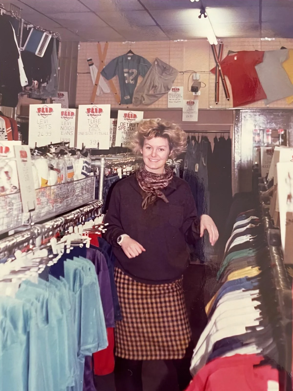 A woman with short blonde hair, wearing a black top, checkered skirt, and scarf, smiling and standing inside a vintage clothing store, surrounded by racks of colorful clothes and signs selling various items.