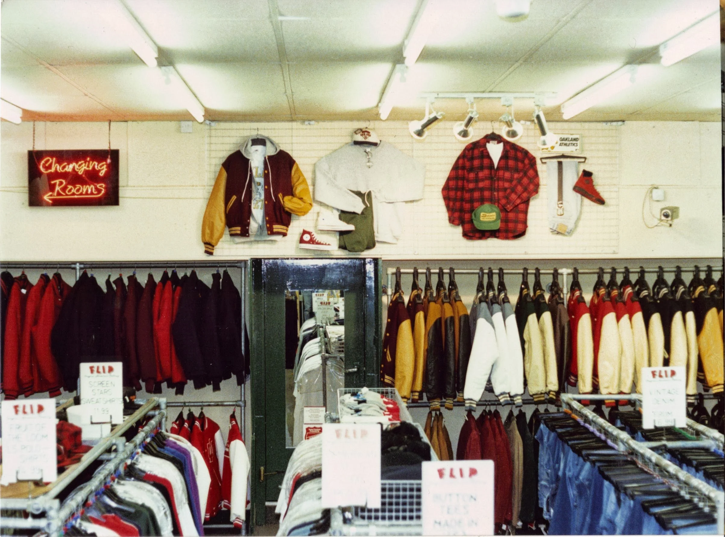 Interior of a vintage clothing store featuring racks of red, white, yellow, and blue jackets and sweaters, with signs indicating sale items and categories, and a wall display of jackets and shirts.