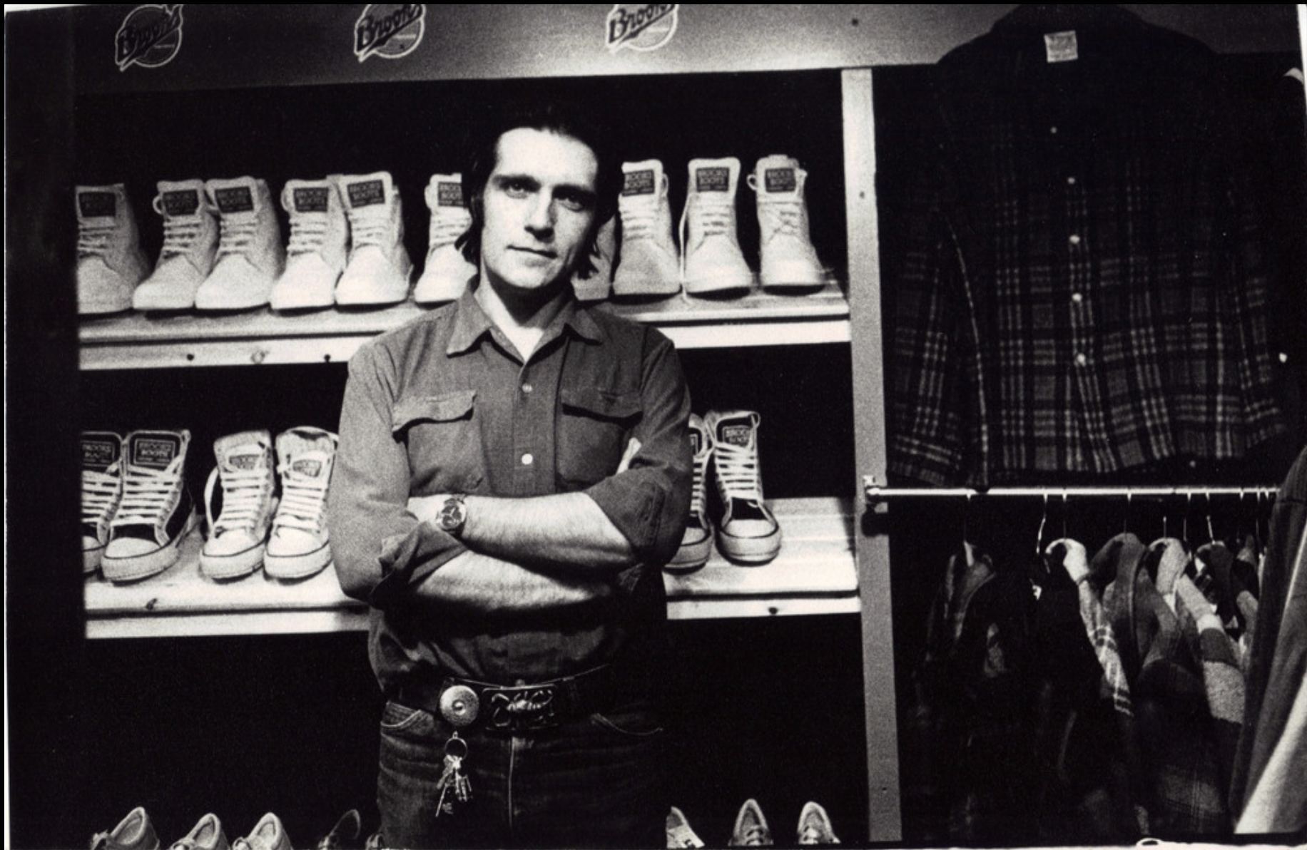 Black and white photo of John Woods standing in a shoe store with his arms crossed, surrounded by shelves of sneakers and boots, and clothing items hanging on racks.