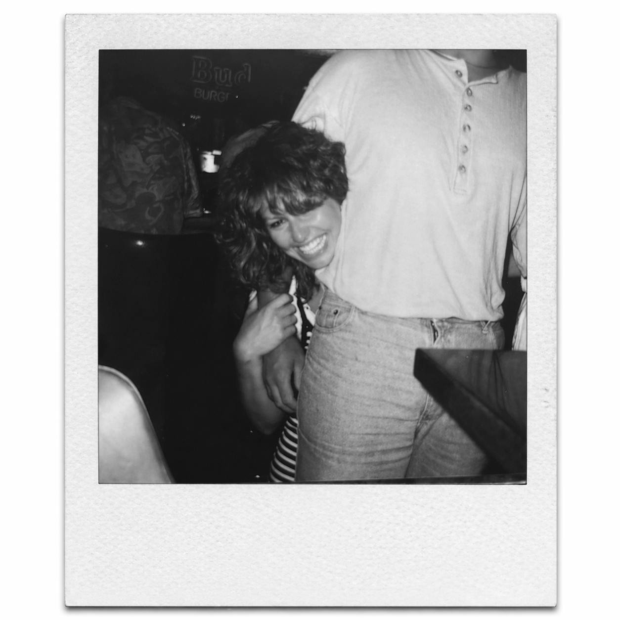 A young woman with curly hair smiling and hugging a person whose face is not visible, inside a dimly lit room with a sign that says 'Bud Burg' in the background.