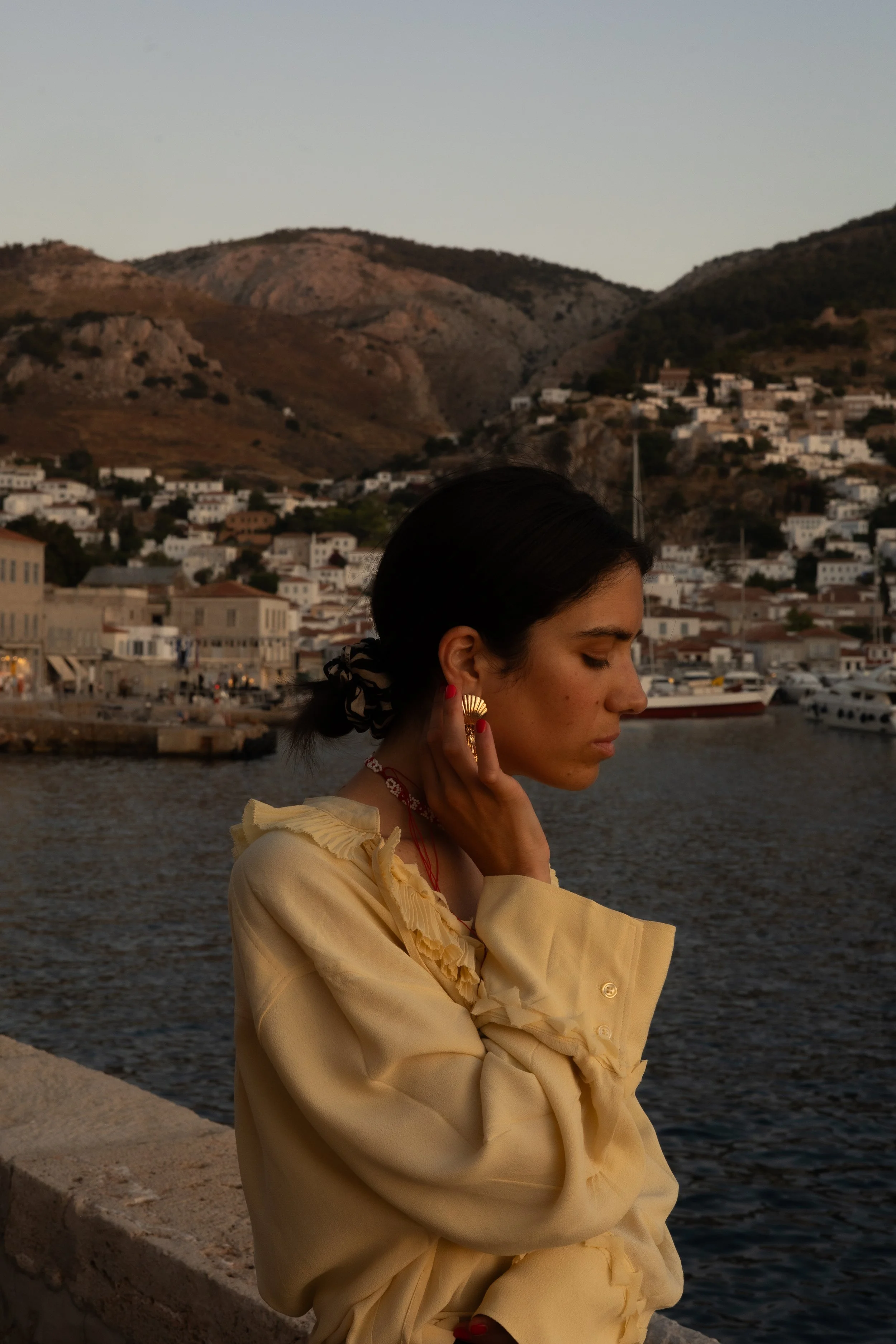 A woman with dark hair and earrings stands by a waterfront, with a town and mountains in the background during dusk.