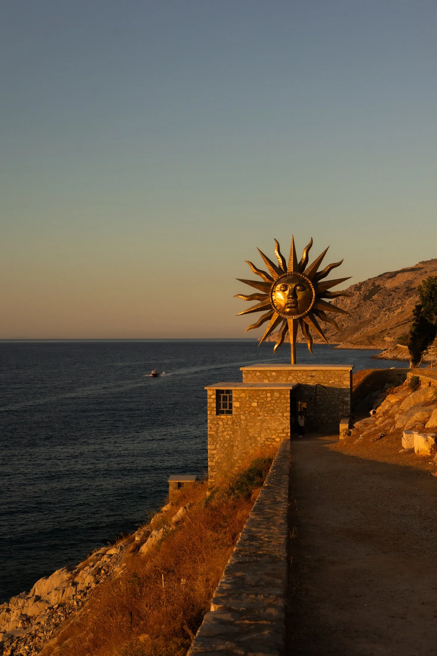 A large decorative sun sculpture with a face, situated on a stone building by the sea at sunset, with hills in the background.