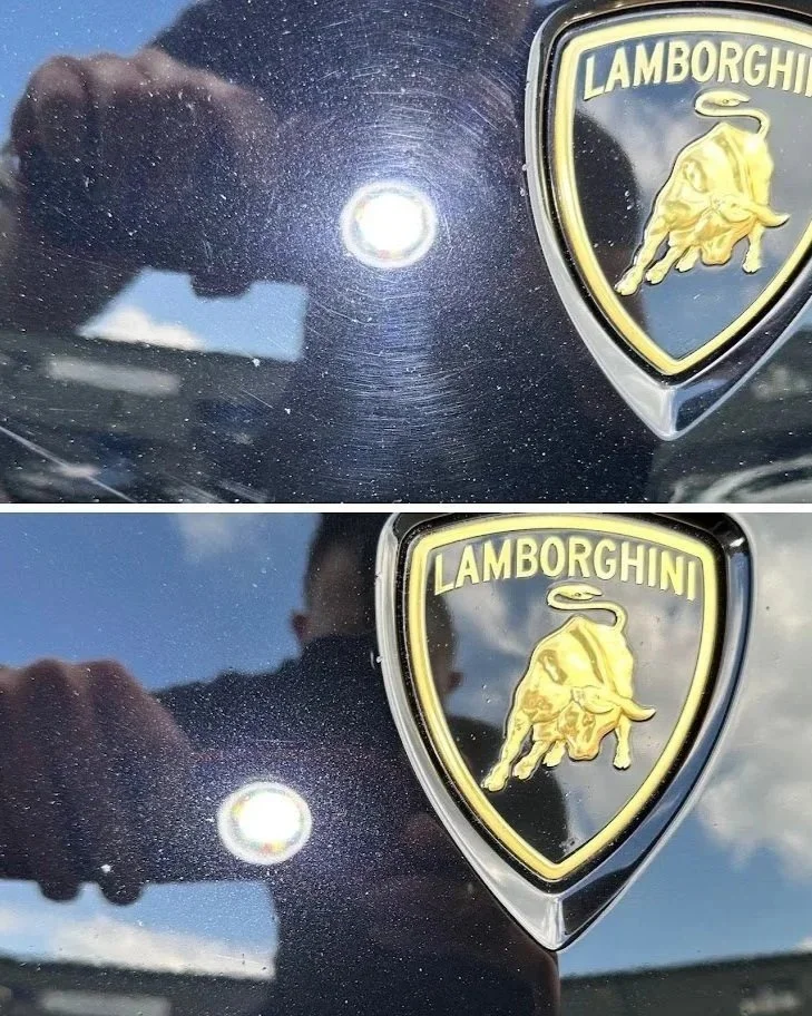 Close-up of a Lamborghini badge on a dark-colored vehicle, reflecting the sky and a person taking the photo.