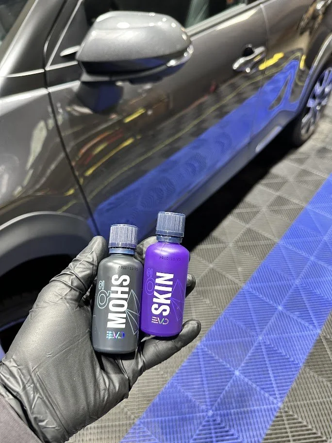 Person wearing a glove holding two bottles labeled MoHS and S-KIN EVO in front of a black car in a garage or workshop.