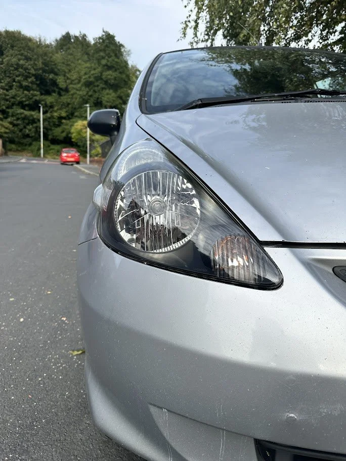 Close-up of the front left side of a silver car, showing the headlight and part of the hood, with a parking lot and trees in the background.