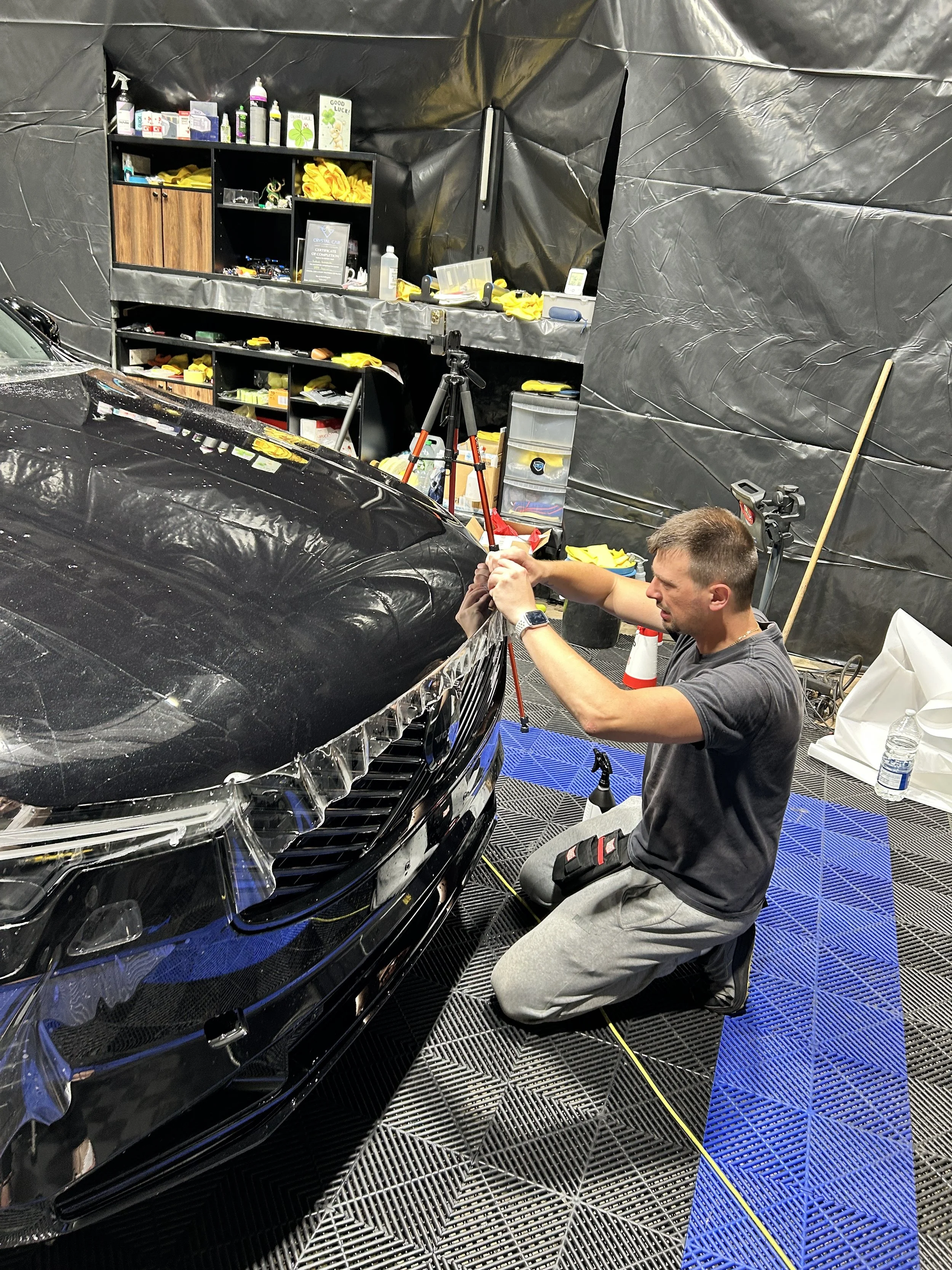 A man kneeling on black and blue checkered flooring working on a black car inside a garage with black plastic walls. Shelves with various bottles, tools, and yellow cloths are visible in the background.