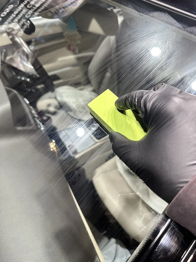 Person wearing a black glove cleaning a car windshield with a yellow scrub pad.