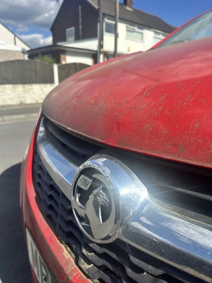 Close-up of a red car with a Vauxhall emblem on the front grille, showing dust and dirt on the hood.