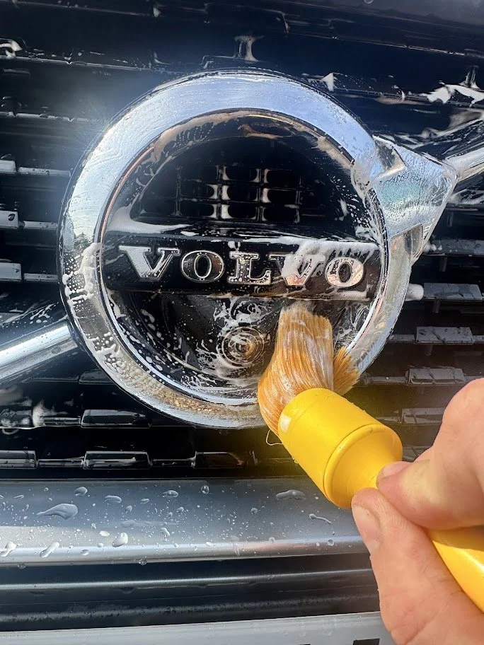 Cleaning a Volvo car emblem with a yellow brush and soapy water.