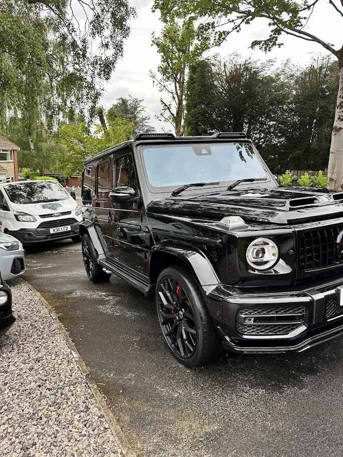 Black Mercedes-Benz G-Class SUV parked on a driveway surrounded by other cars and trees.