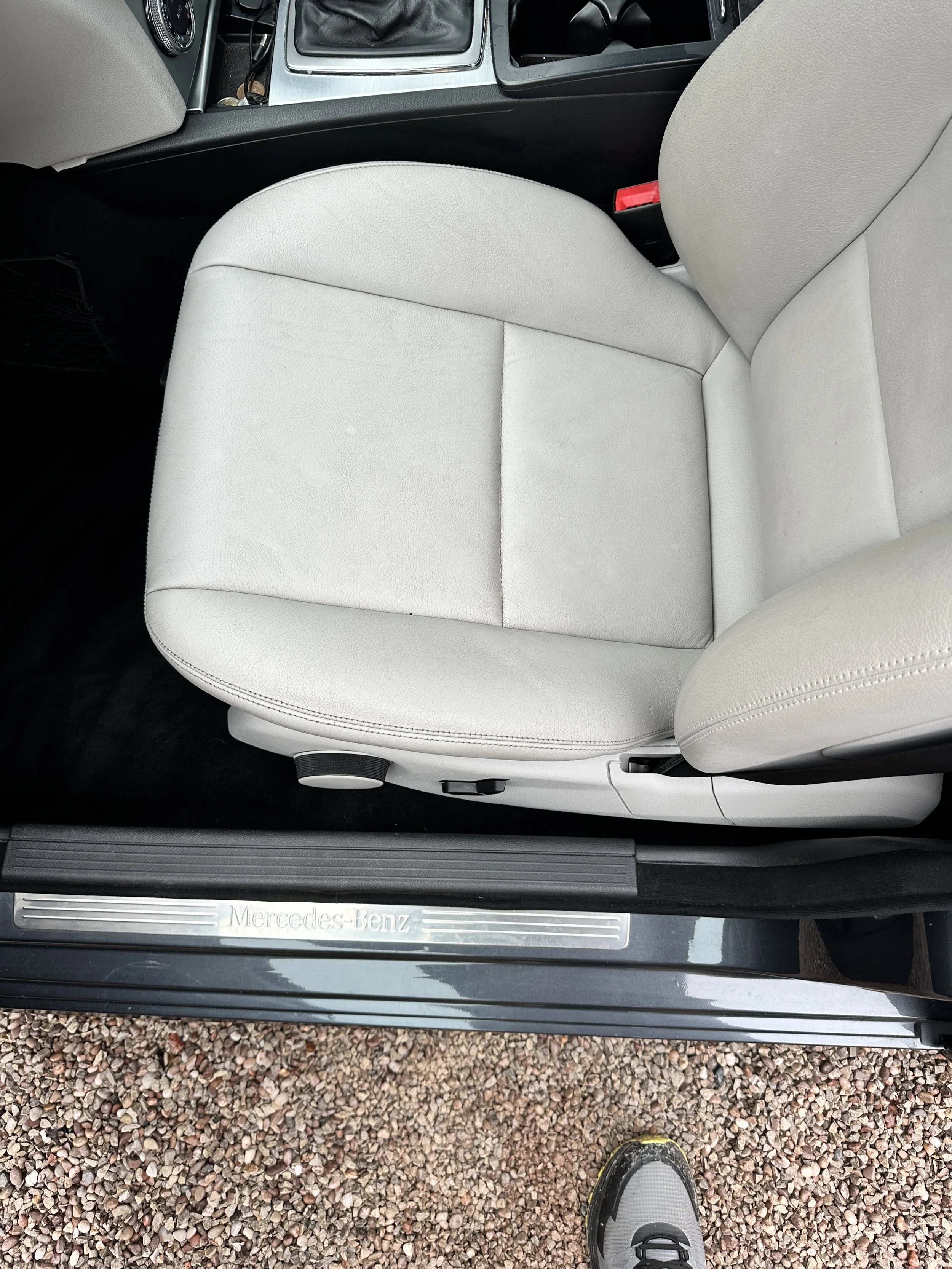 Inside a car showing a light-colored leather passenger seat, door sill with Mercedes-Benz logo, and part of the floor covered with small gravel stones.
