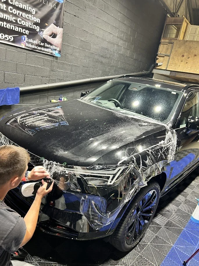 A black and blue car inside a garage, having clear protective film applied to its front end by a person using a spray bottle and a squeegee. The garage has a gray brick wall and a banner advertising car detailing services and ppf