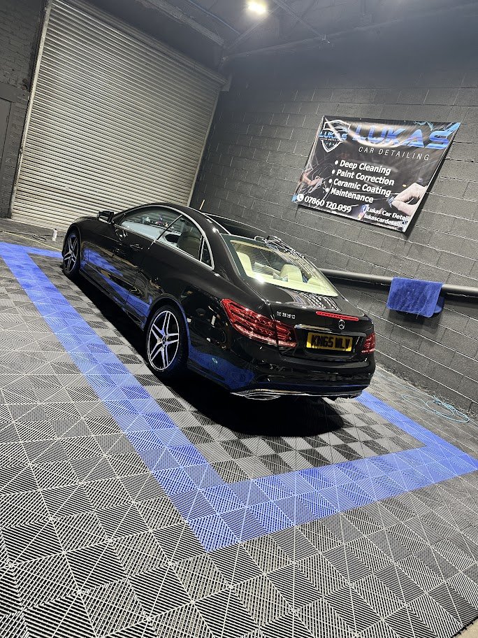 Black luxury car parked inside a detailing shop with a blue advertising banner on the wall and a blue towel hanging on the wall.