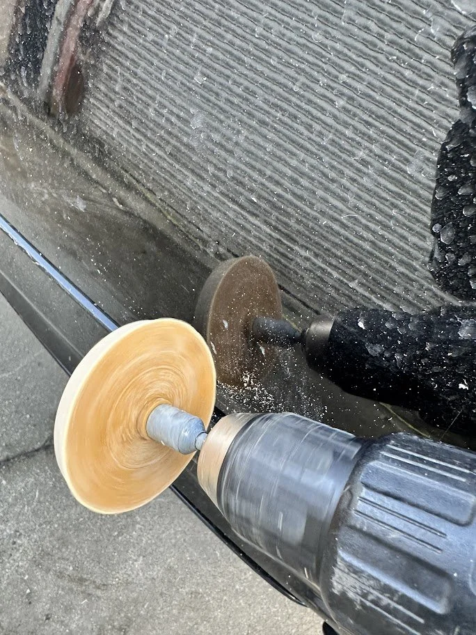 A person using a power grinder with a sanding disc on a black vehicle's surface.