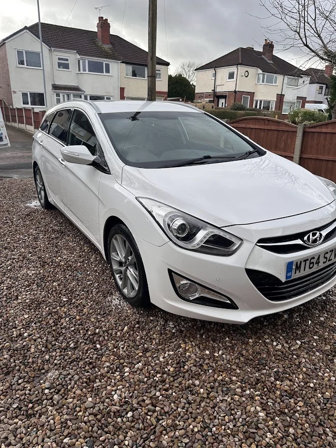 A white Hyundai sedan parked on a gravel driveway in front of residential houses with gardens and fences.