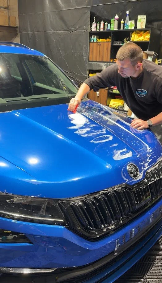 A man is cleaning the hood of a blue car with a red cloth inside a garage or workshop. The workshop has shelves with cleaning supplies PPF foil application - lukas car detailing - manchester