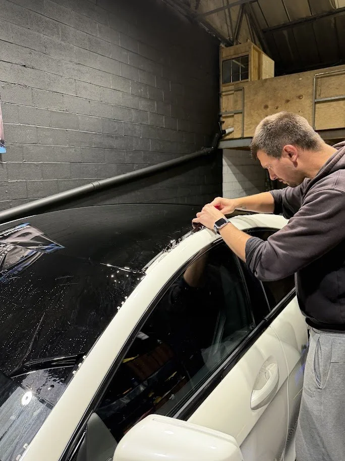 A man installing a decal or adhesive wrap on the roof of a white sports car with a black roof, in a garage or workshop setting.