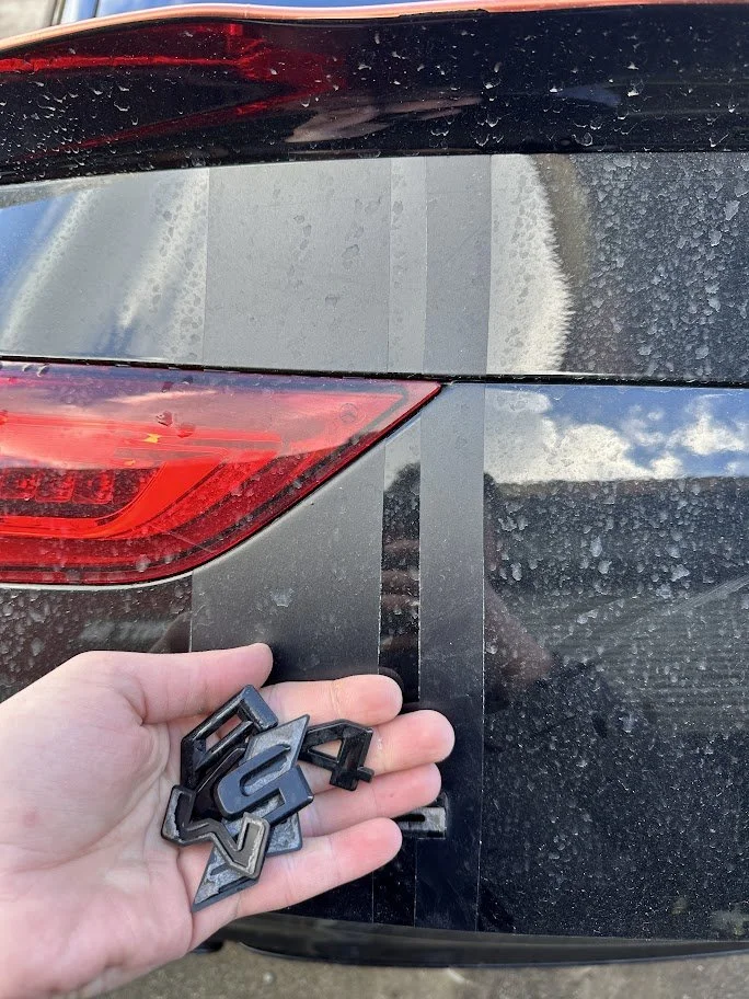 Person holding black plastic badges or emblems with the number '4' in front of a black car with a red tail light and people reflected on its surface.