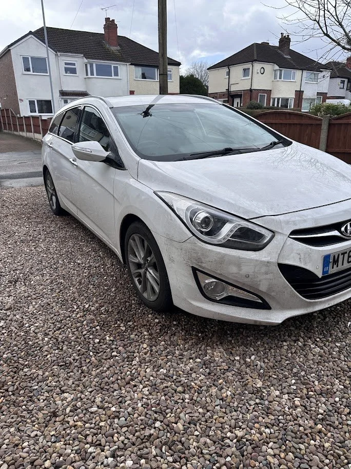 A white Hyundai hatchback car parked on a gravel driveway in a residential neighborhood with houses in the background.