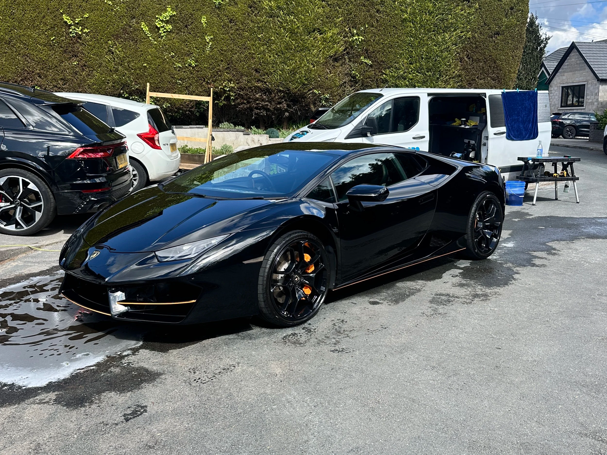 A black luxury sports car is parked on a wet driveway, with other cars and a work table with tools nearby.
