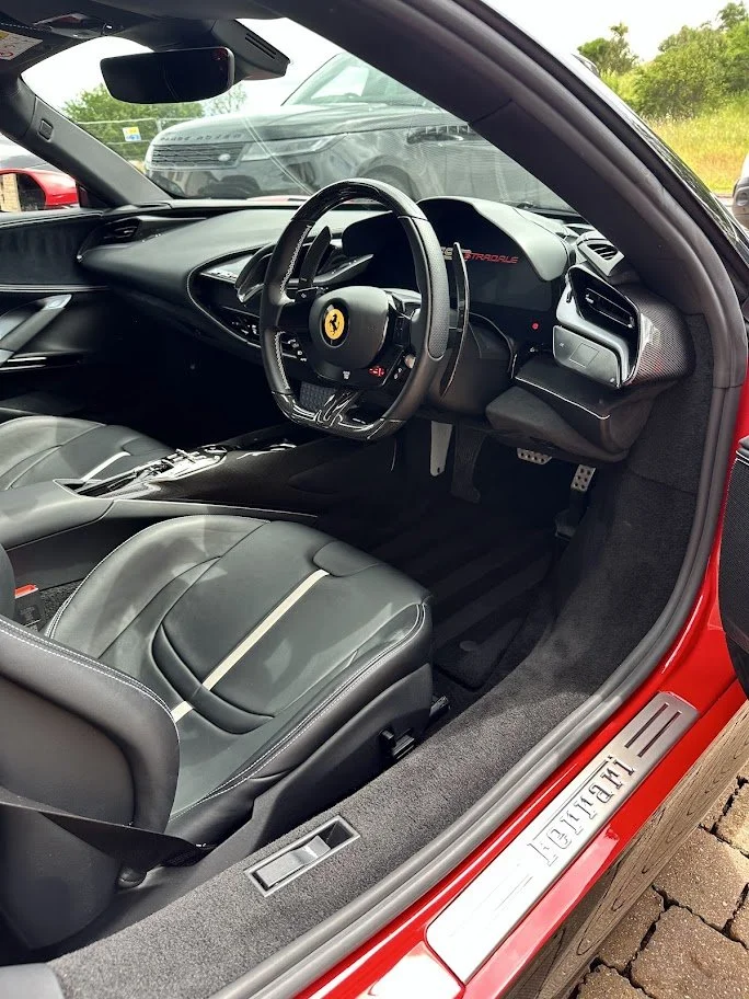 The interior of a red Ferrari sports car, showing the leather seat, steering wheel with Ferrari logo, dashboard with digital display, and part of the door sill with 'Ferrari' branding.