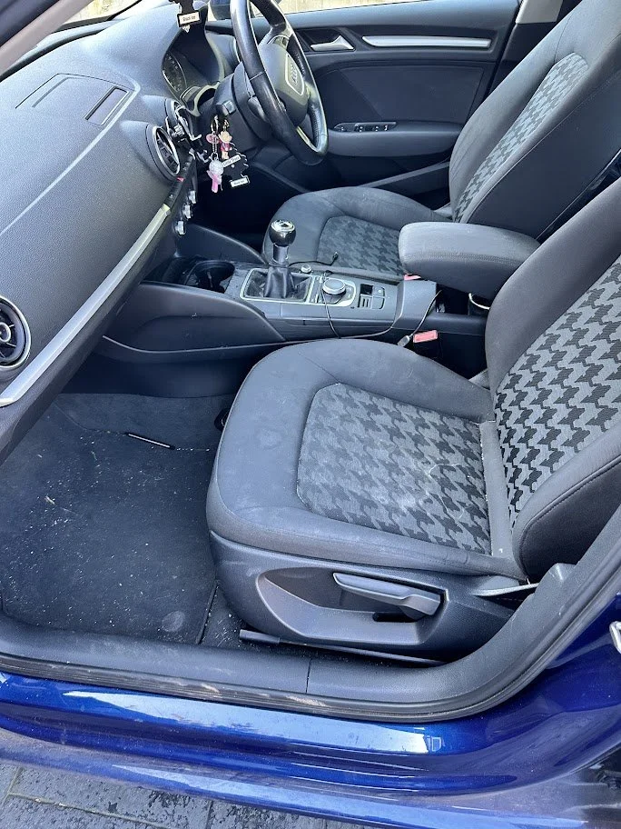 Interior of a car showing the driver's seat and front passenger seat, gear shift, steering wheel with keys hanging, and dashboard with some hanging accessories.