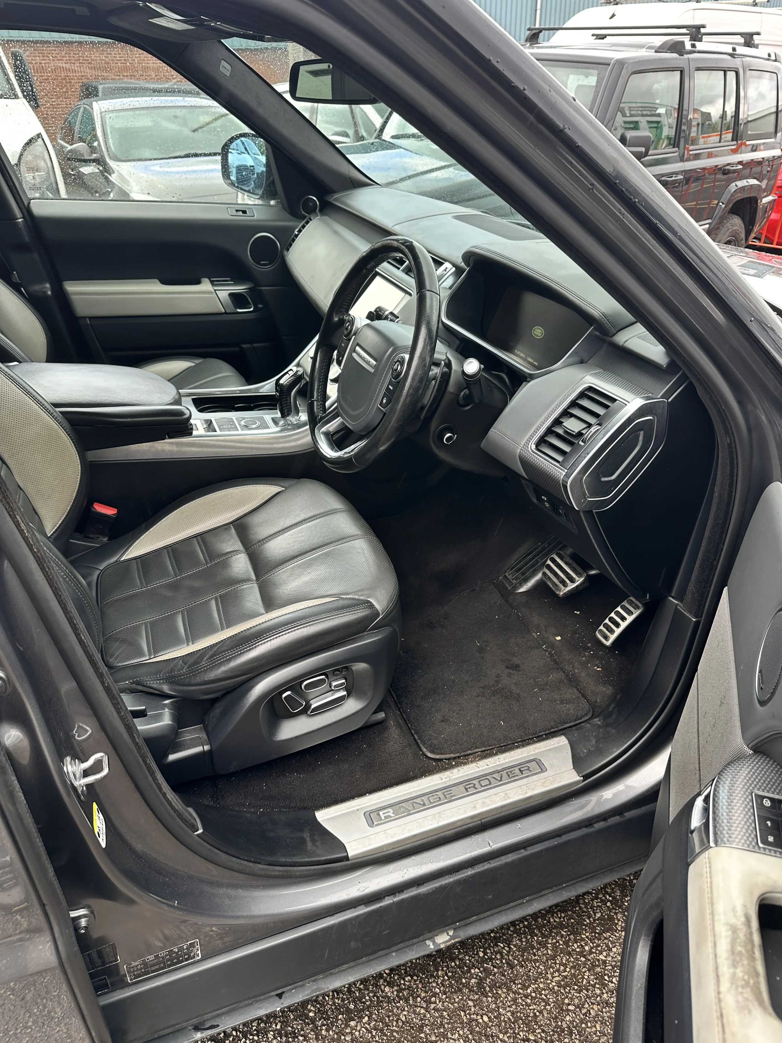 The interior of a Range Rover vehicle with black leather seats, a steering wheel, and dashboard, parked among other cars on a rainy day.