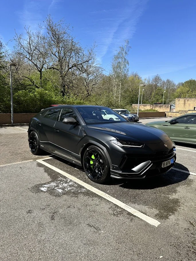 A black luxury coupe car parked in a parking lot with trees and blue sky in the background.