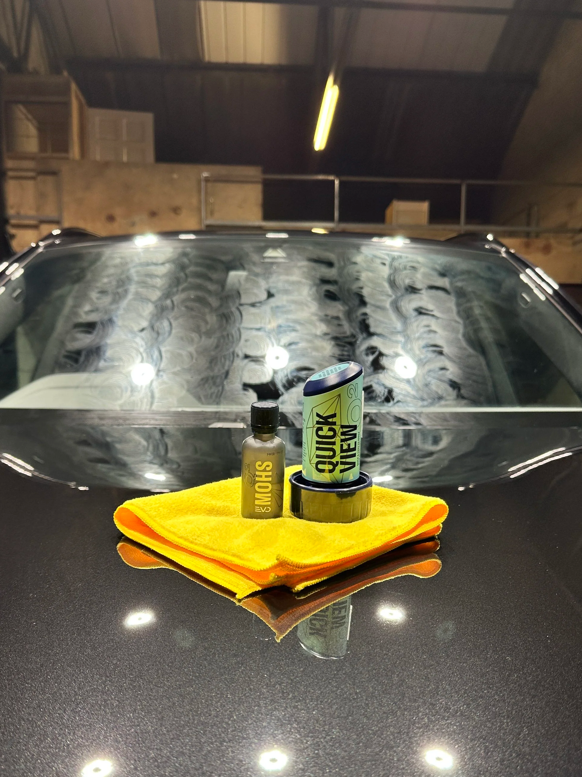 A display of car detailing products, including a spray bottle, a container of quick wax, and a yellow microfiber cloth, on a black reflective surface in a garage with wooden walls and overhead lighting.