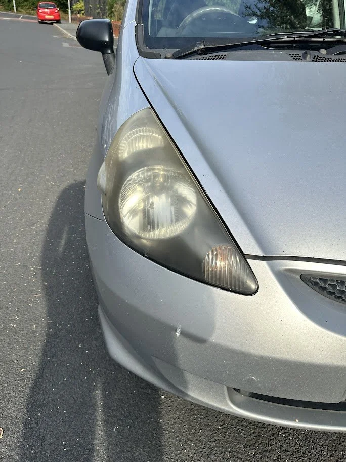 Close-up of the front left side of a silver hatchback car showing a foggy headlight and a small dent on the bumper, parked on an asphalt street.