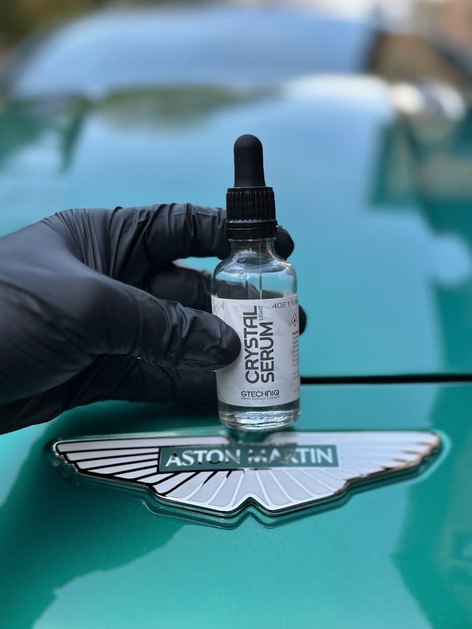 A hand wearing a black glove holds a small bottle of Crystal Serum beneath the Aston Martin logo on a turquoise car.