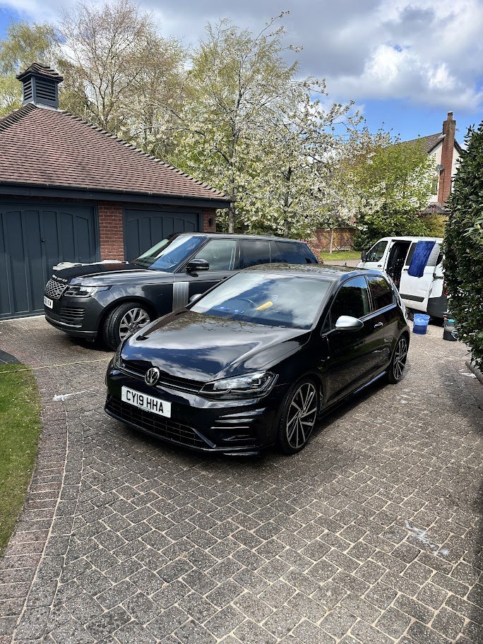 A black Volkswagen car parked on a brick driveway in front of a garage, with other vehicles and trees in the background.