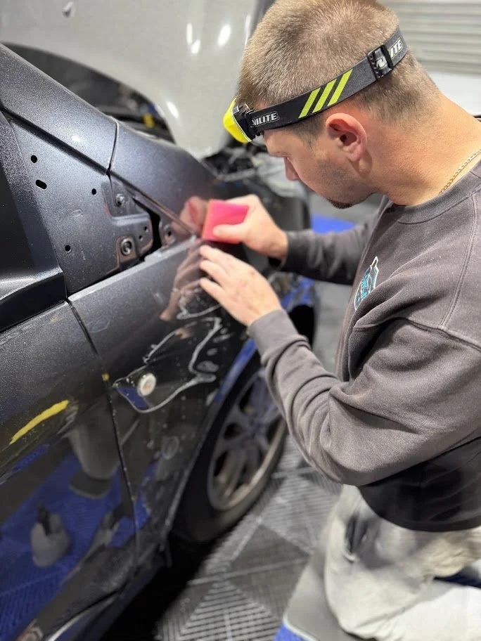 A man wearing a headlamp is repairing a black car, using a small pink tool on the side of the vehicle inside a workshop.