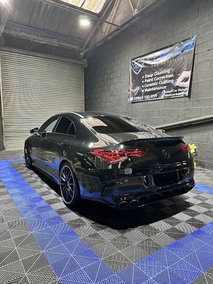 Black Mercedes-Benz sedan parked indoors at an auto detailing shop with a banner on the wall listing services like deep cleaning, paint correction, ceramic coating, and maintenance.