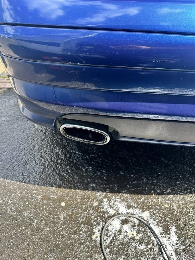 Close-up of the rear of a blue car showing the exhaust pipe and damaged paint on the bumper, with a wet driveway and some spilled powder or debris on the ground.