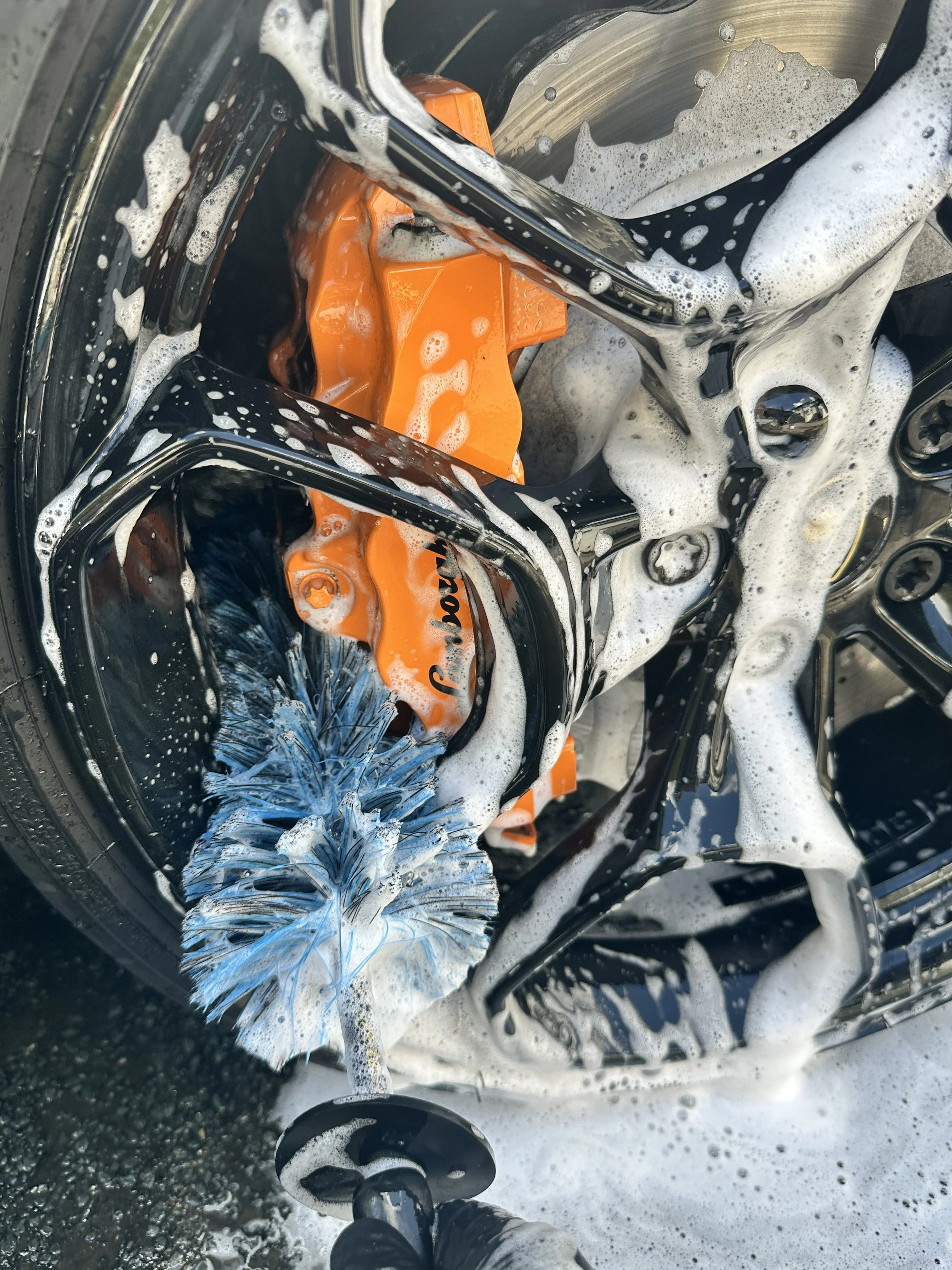 View of a car wheel being cleaned with a blue brush, soap suds covering the wheel, and a person's feet visible at the bottom of the image.