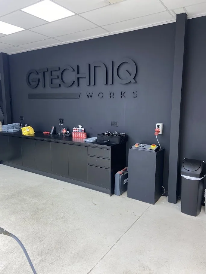 Office break area with black cabinetry, supplies, and a large wall sign reading 'GTECHIQ WORKS' in black letters against a dark gray wall.