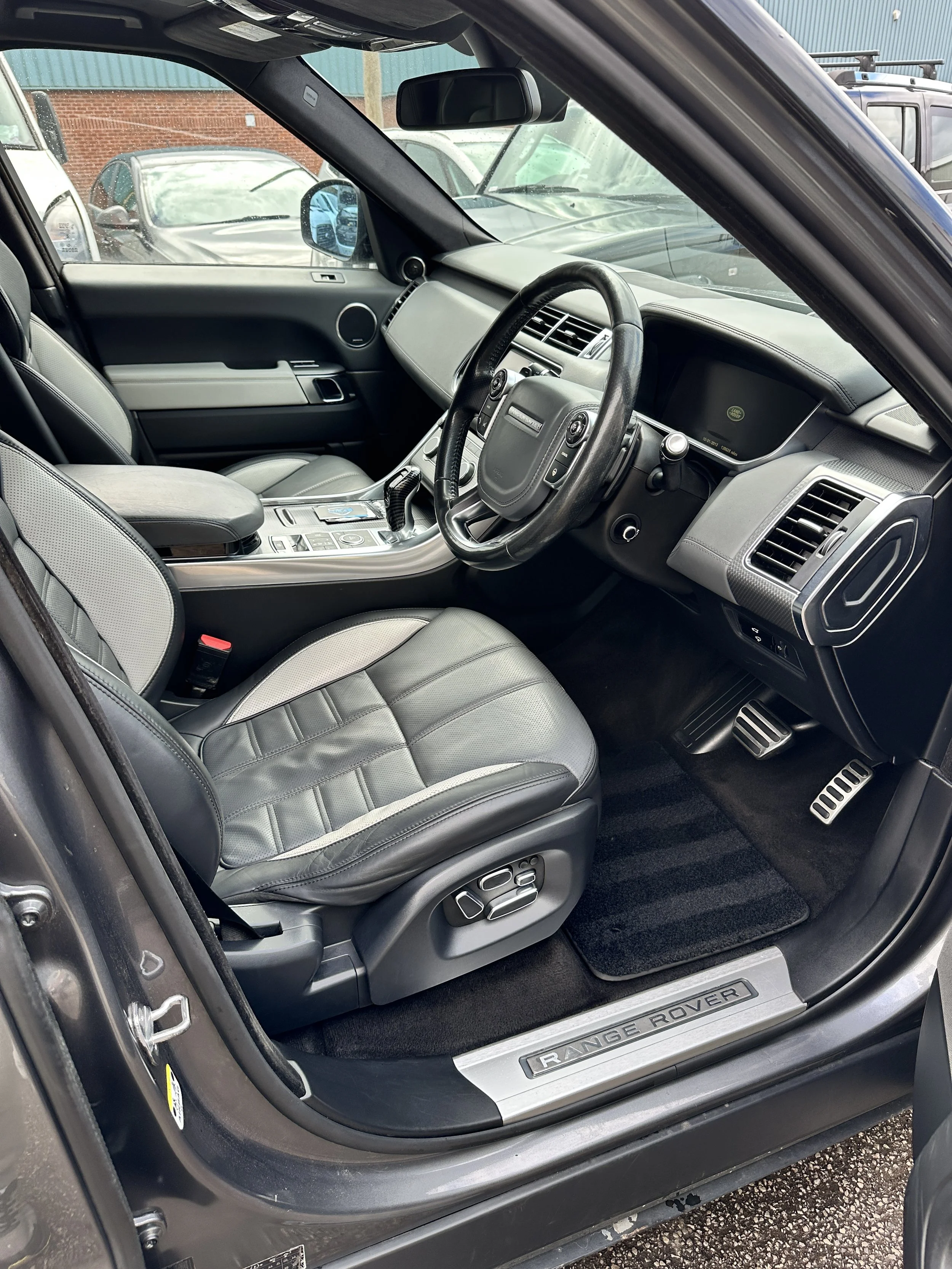 Interior of a Land Rover vehicle showing the driver and passenger seats, steering wheel, center console, and dashboard, with a view through the windshield of parked cars and a brick wall.