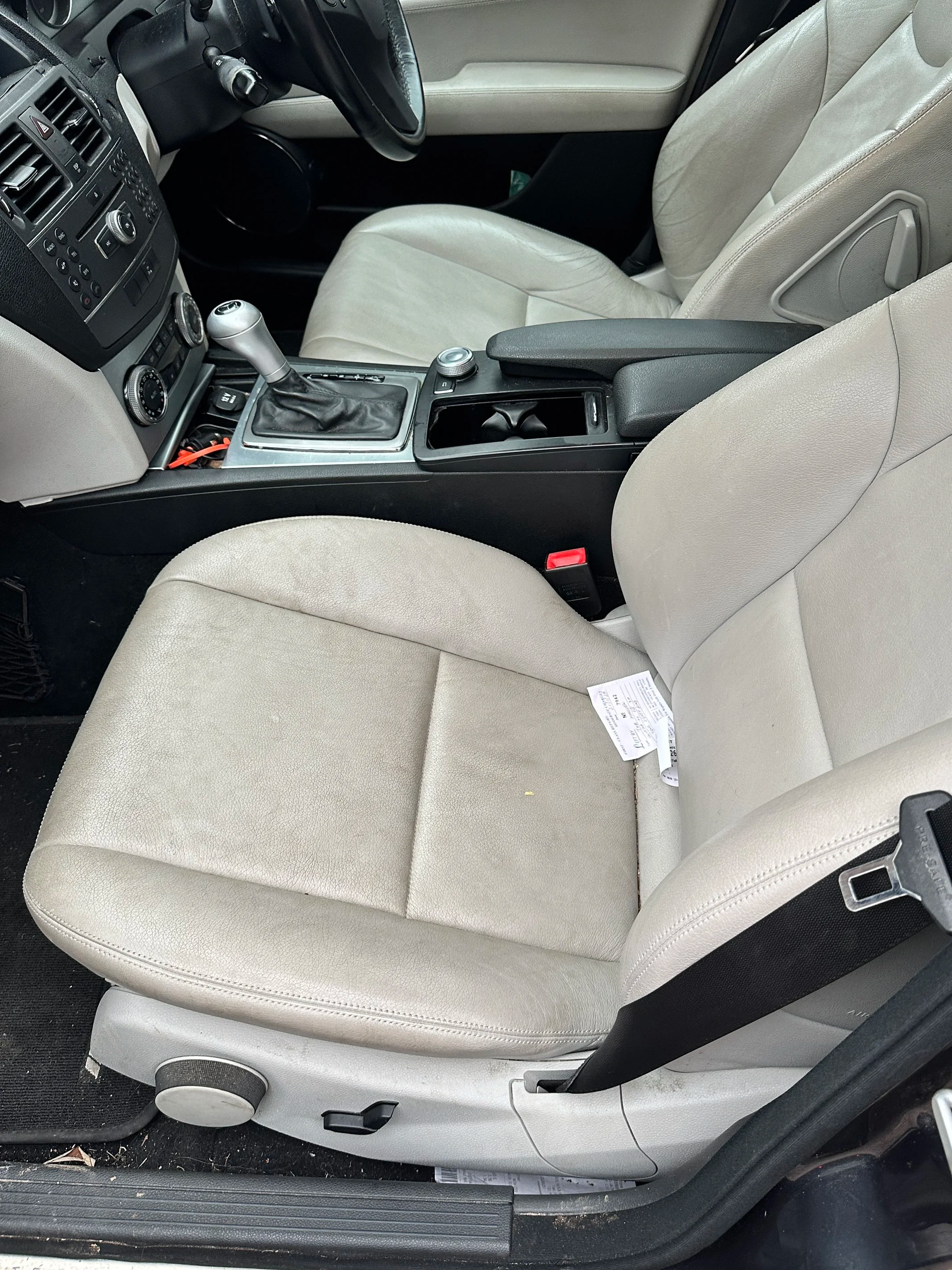 The image shows the interior of a car, featuring beige seats, a center console with a gear shift, buttons, and a handbrake. The floor and some areas near the door show signs of dirt.