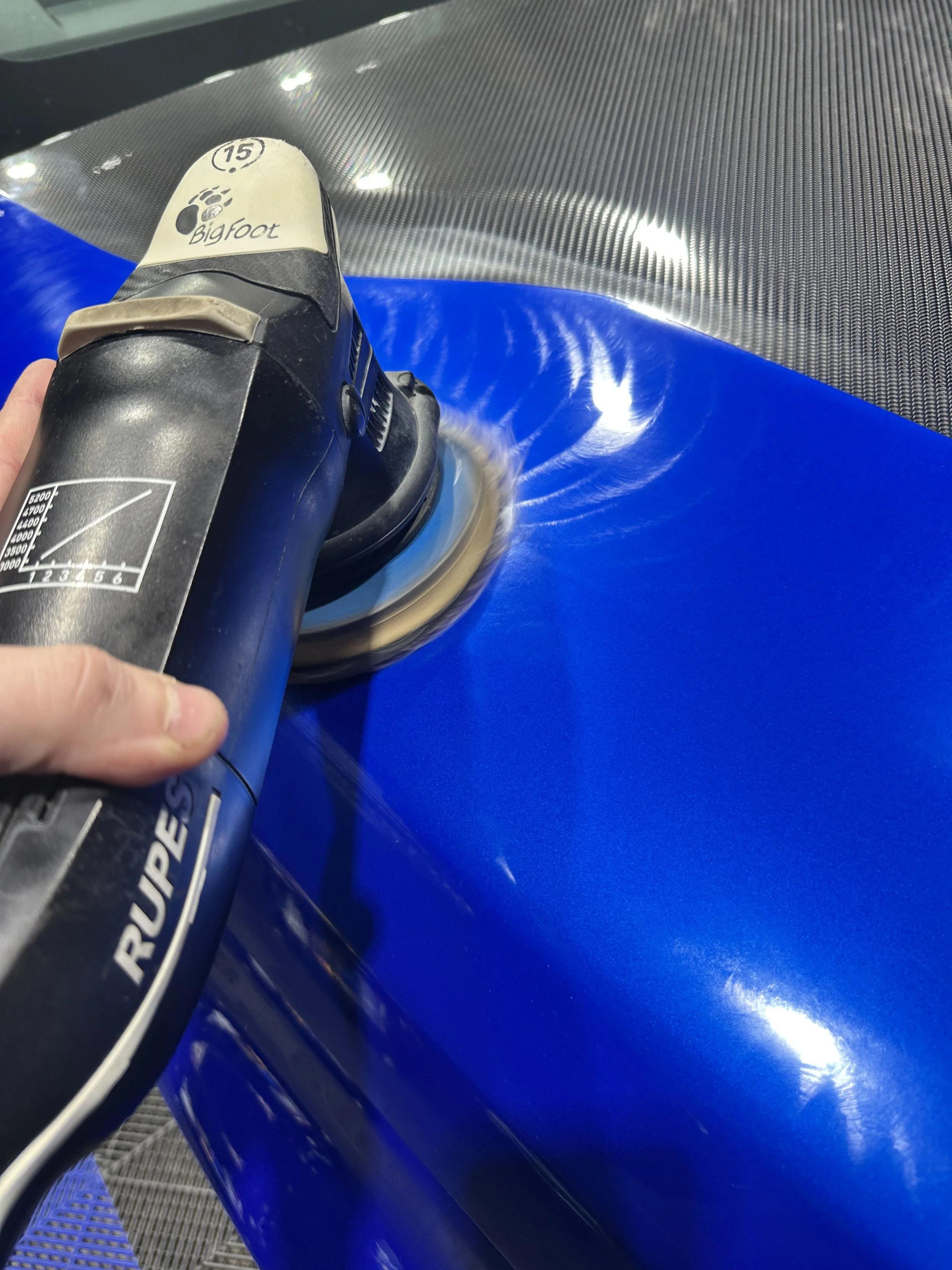 A person holding a black aerosol spray can labeled with temperature and valve information, spray coming out onto a blue surface, near a textured metallic surface and a plastic grate.