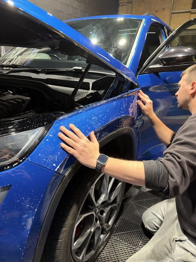 A man inspecting the fender of a blue SUV with an open hood, inside a garage. PPF foil application - lukas car detailing - manchester
