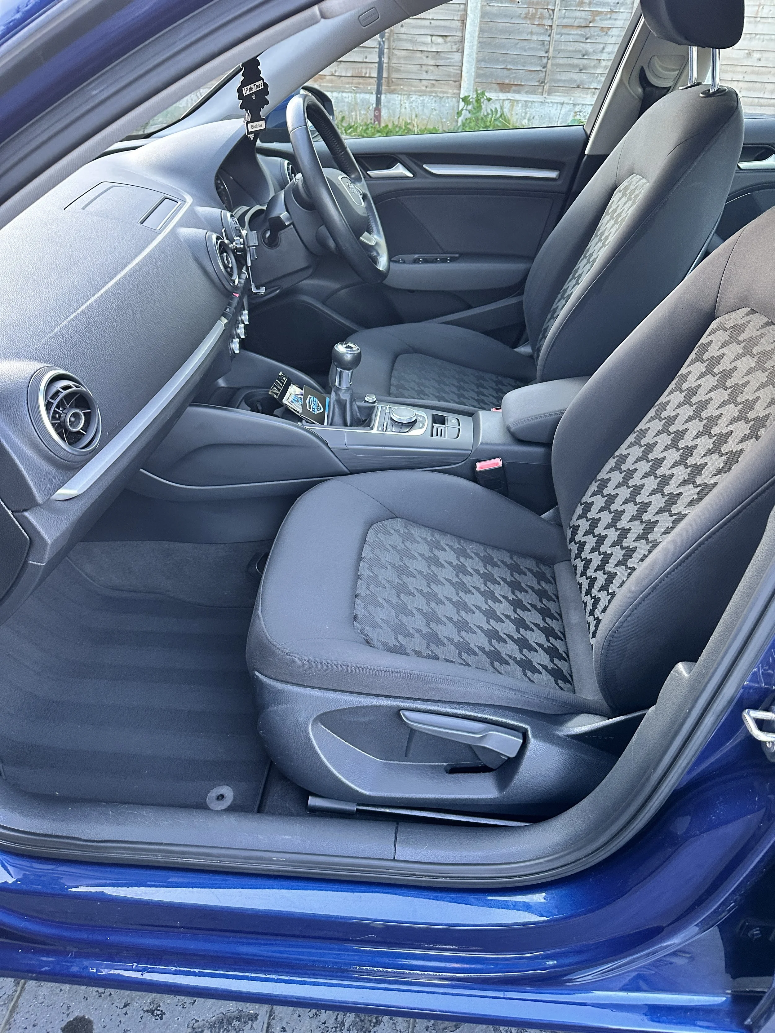 The interior of a blue car showing the front passenger and driver seats, dashboard, steering wheel, gear shift, and center console, with a wooden fence and some greenery outside the window.
