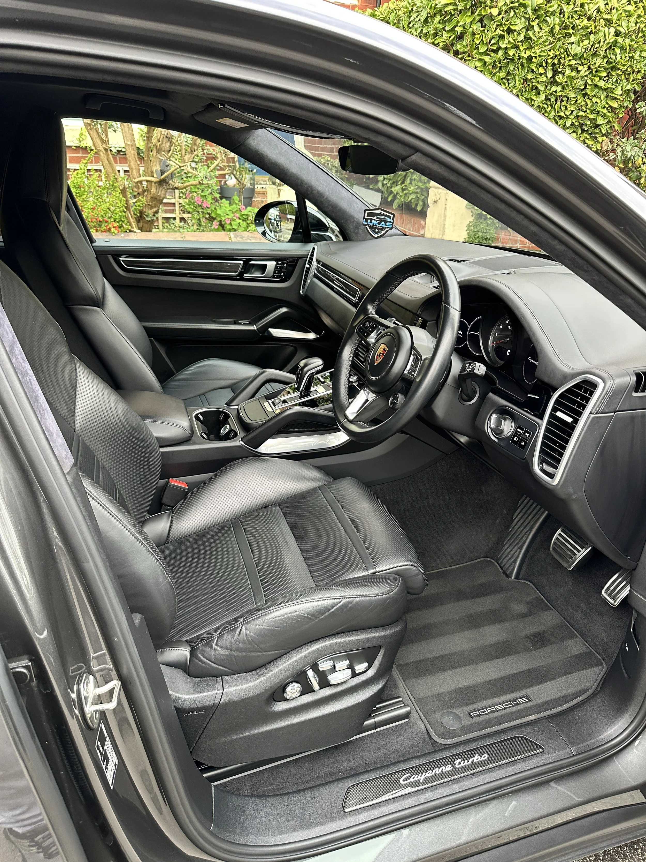 Interior of a luxury car featuring black leather seats, a modern dashboard, and Porsche branding on the floor mat, with a view of the garden outside the vehicle.
