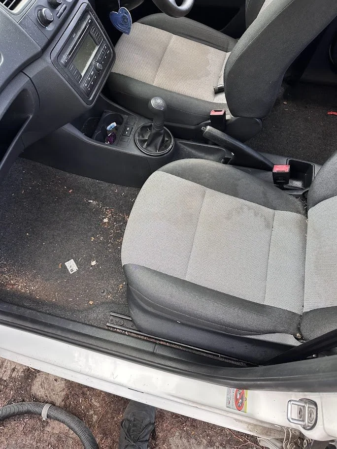 Interior of a car showing the driver's seat, passenger seat, gear shift, handbrake, and dashboard, with some dirt and debris on the floor.