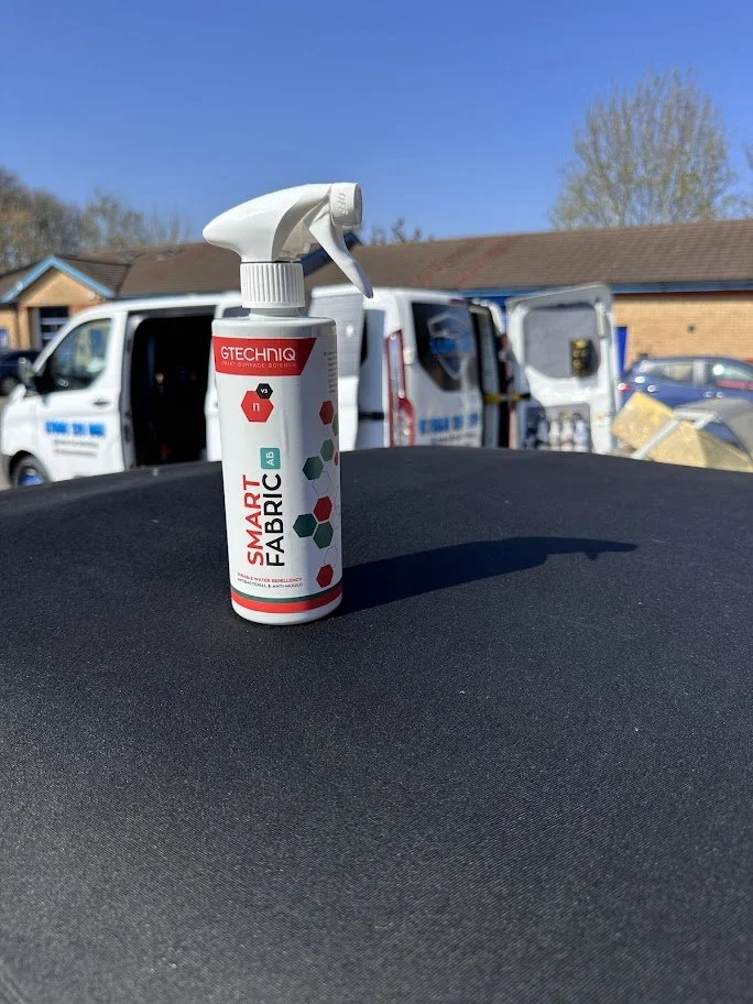 A spray bottle labeled 'Smart Fabric' in the foreground, with a background of service vans and a building under a clear blue sky.