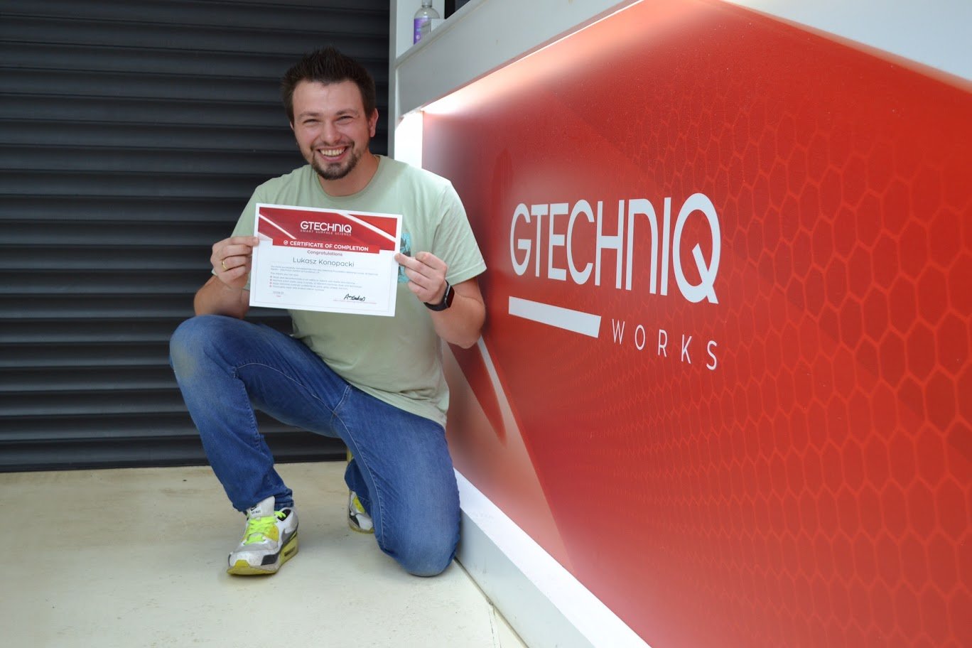 Man holding a certificate and smiling, kneeling next to a large red sign that says 'GTECH IQ WORKS'.