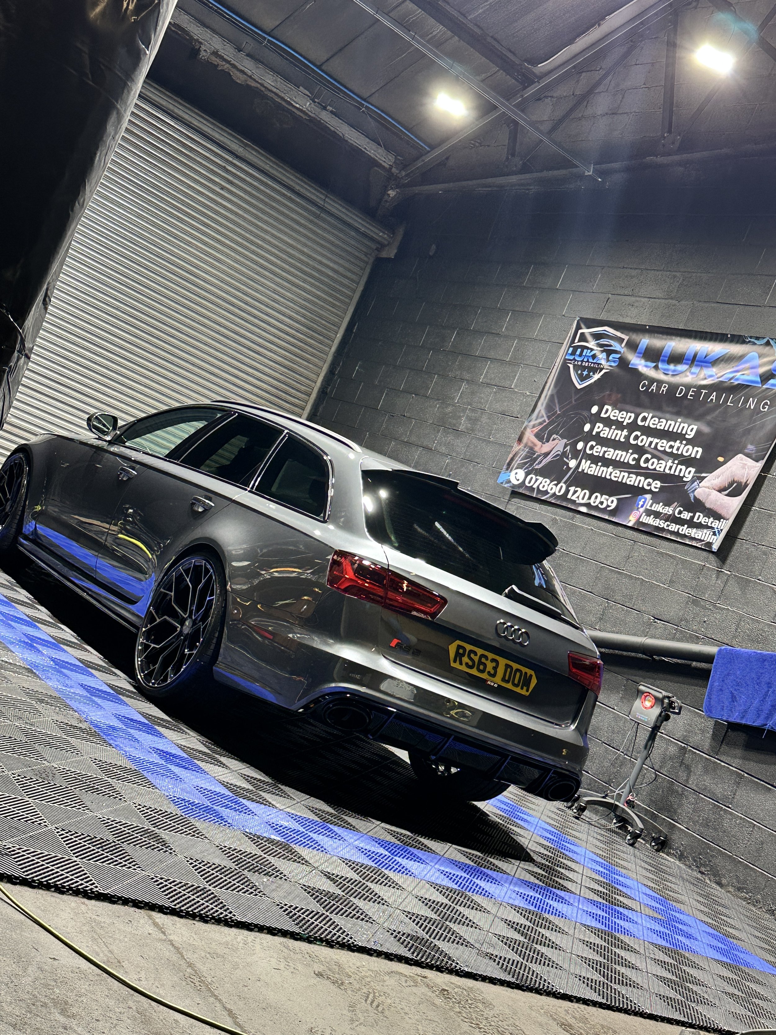 A silver Audi RS3 parked inside a car detailing shop with a black wall background and a sign that reads 'Lukas Car Detailing' offering various services like deep cleaning and paint correction.