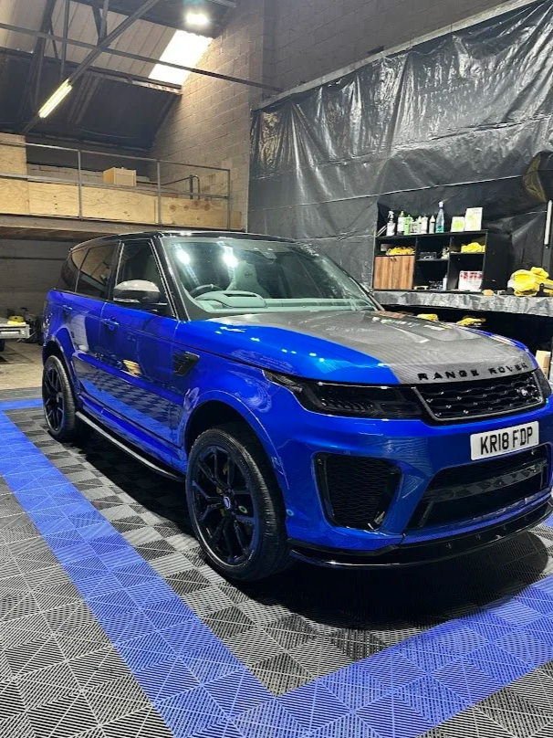 A blue Range Rover SUV with black accents parked inside a garage or workshop, with shelves and cleaning supplies in the background.